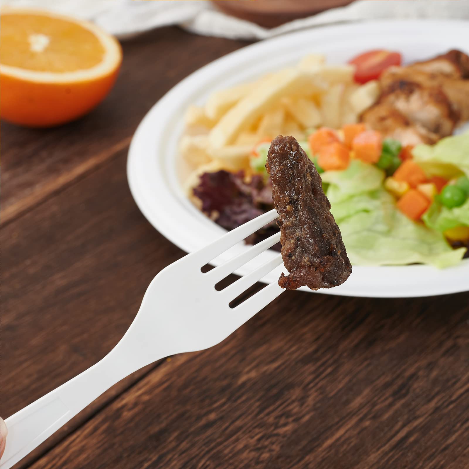 A white fork holds a piece of meat above a colorful plate with salad, fries, and chicken, beside a half orange on a wooden table.