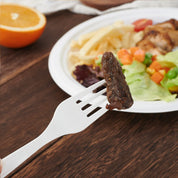 A white fork holds a piece of meat above a colorful plate with salad, fries, and chicken, beside a half orange on a wooden table.