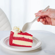 A hand holds a spoon poised to take a bite from a slice of red velvet cake topped with whipped cream on a plate.