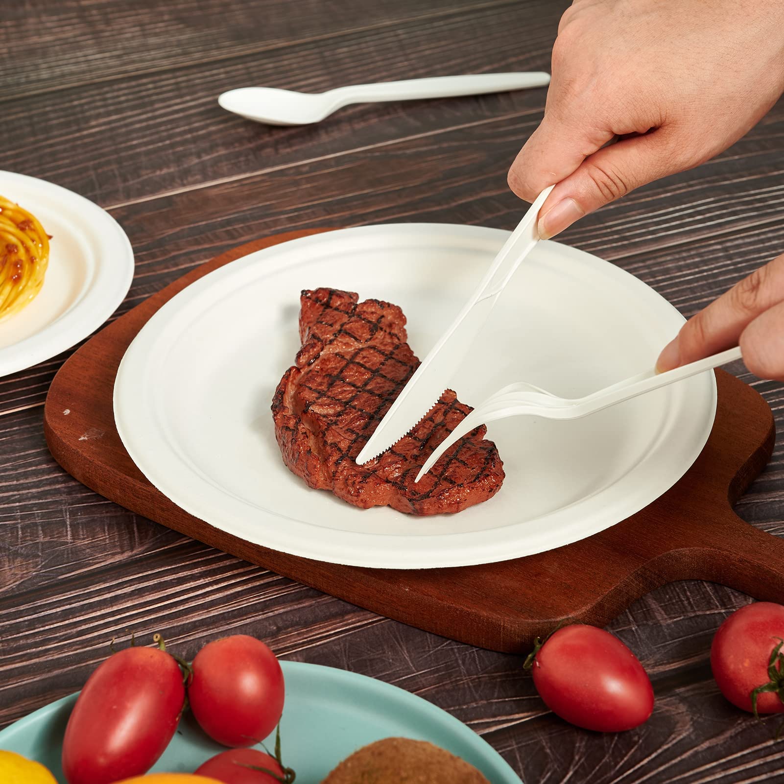 A hand uses white utensils to cut a grilled steak on a ECOLipak white plate, with cherry tomatoes and a pasta dish in the background.