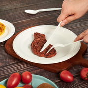 A hand uses white utensils to cut a grilled steak on a ECOLipak white plate, with cherry tomatoes and a pasta dish in the background.