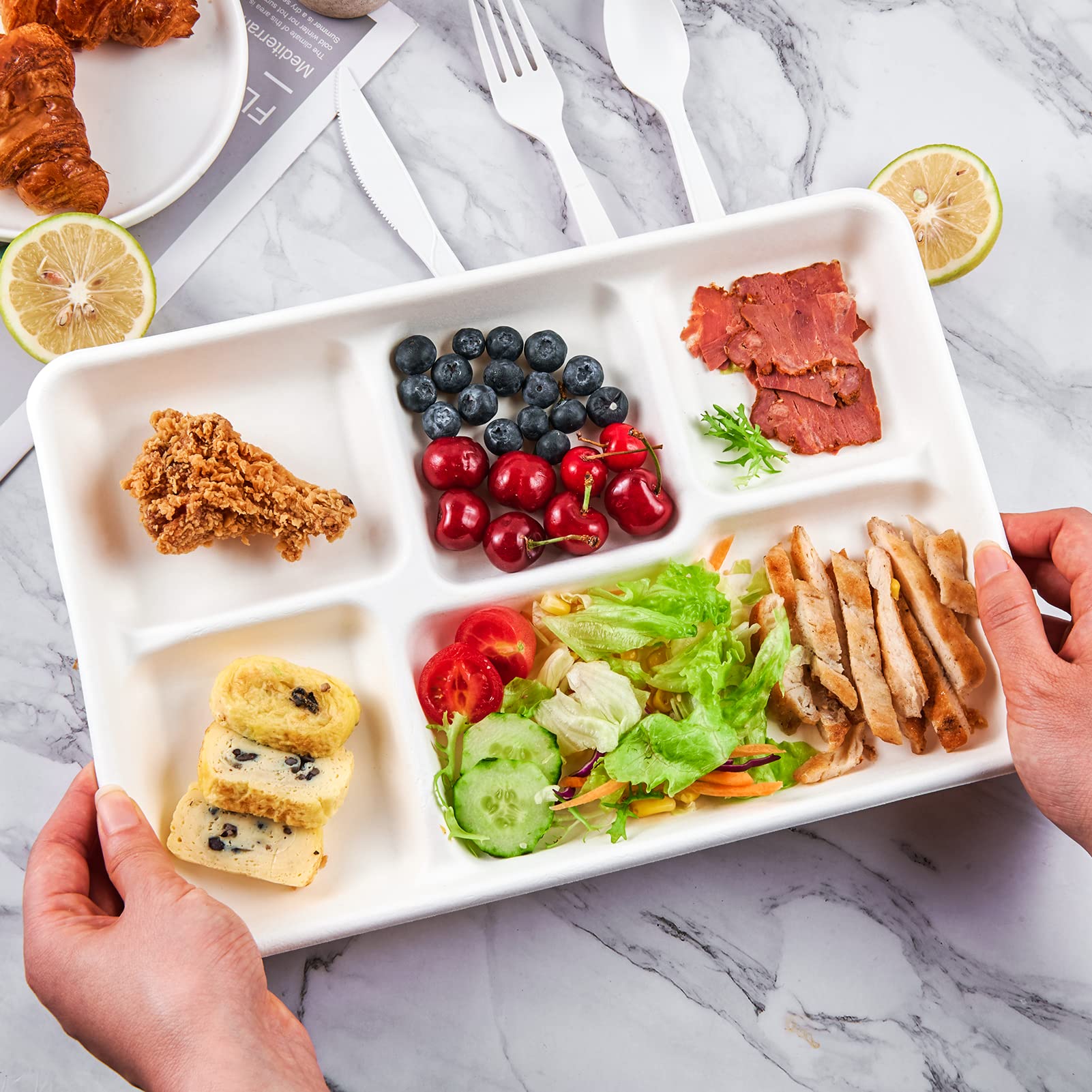 A hand holds a divided food tray with fried chicken, berries, salad, sliced meat, and a dessert, set against a marble background.