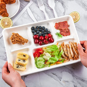 A hand holds a divided food tray with fried chicken, berries, salad, sliced meat, and a dessert, set against a marble background.