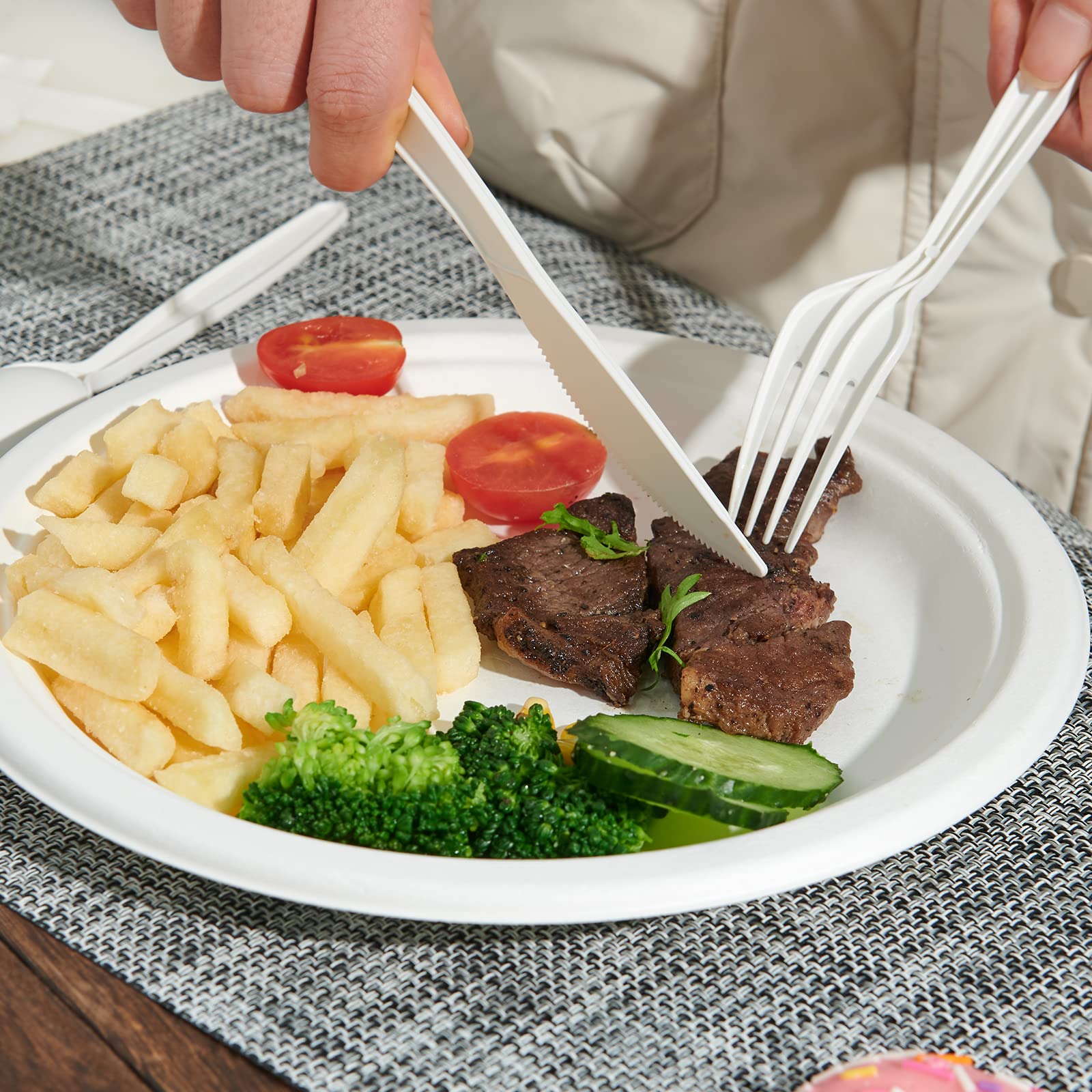 A close-up of a hand cutting a steak on a plate with fries, broccoli, cherry tomatoes, and cucumber, using plastic utensils.
