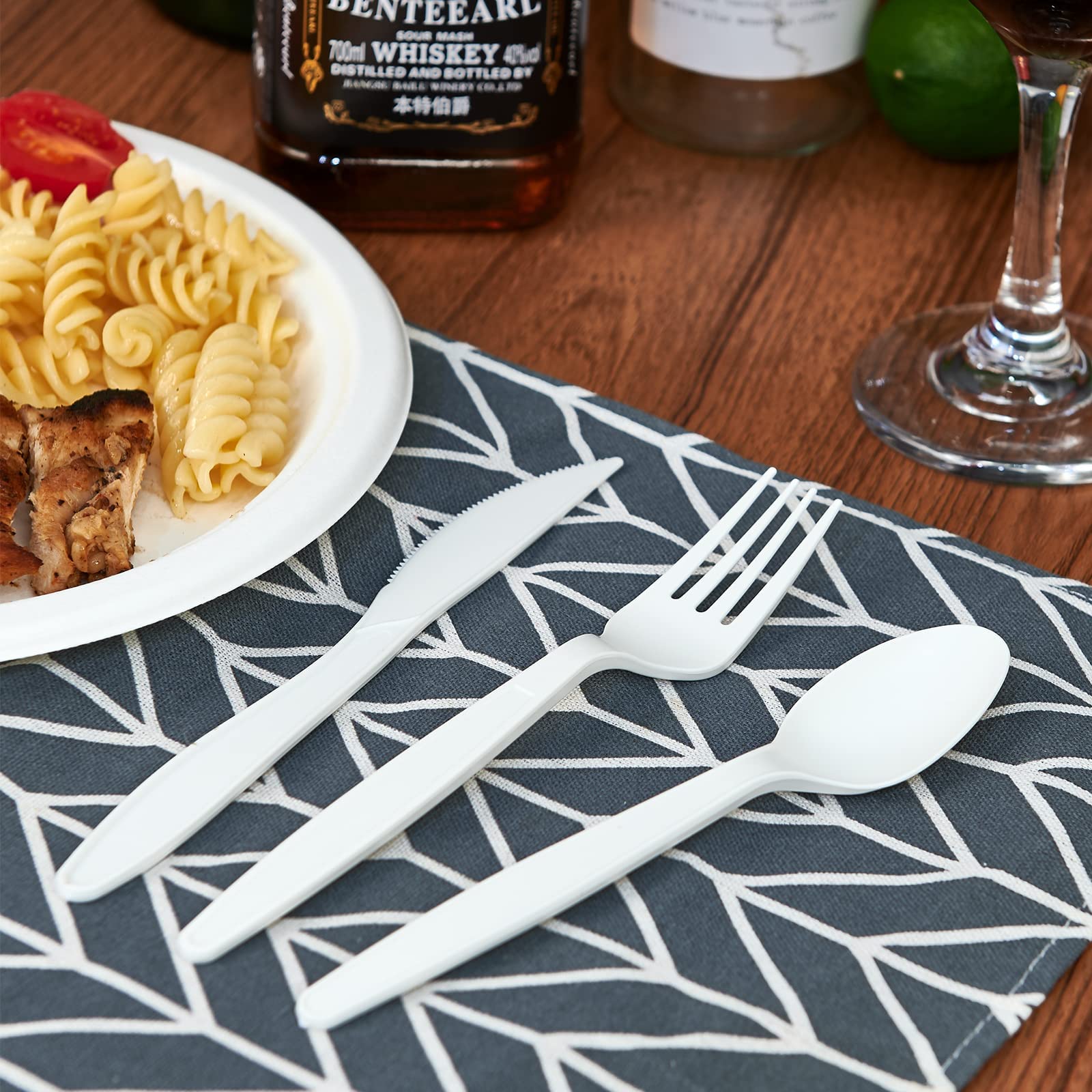 A white plastic knife, fork, and spoon are arranged beside a plate of pasta and grilled chicken on a patterned napkin.