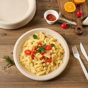 A beige plate of spiral pasta topped with tomato sauce, cherry tomatoes, and mint, surrounded by utensils and a small cup of sauce.