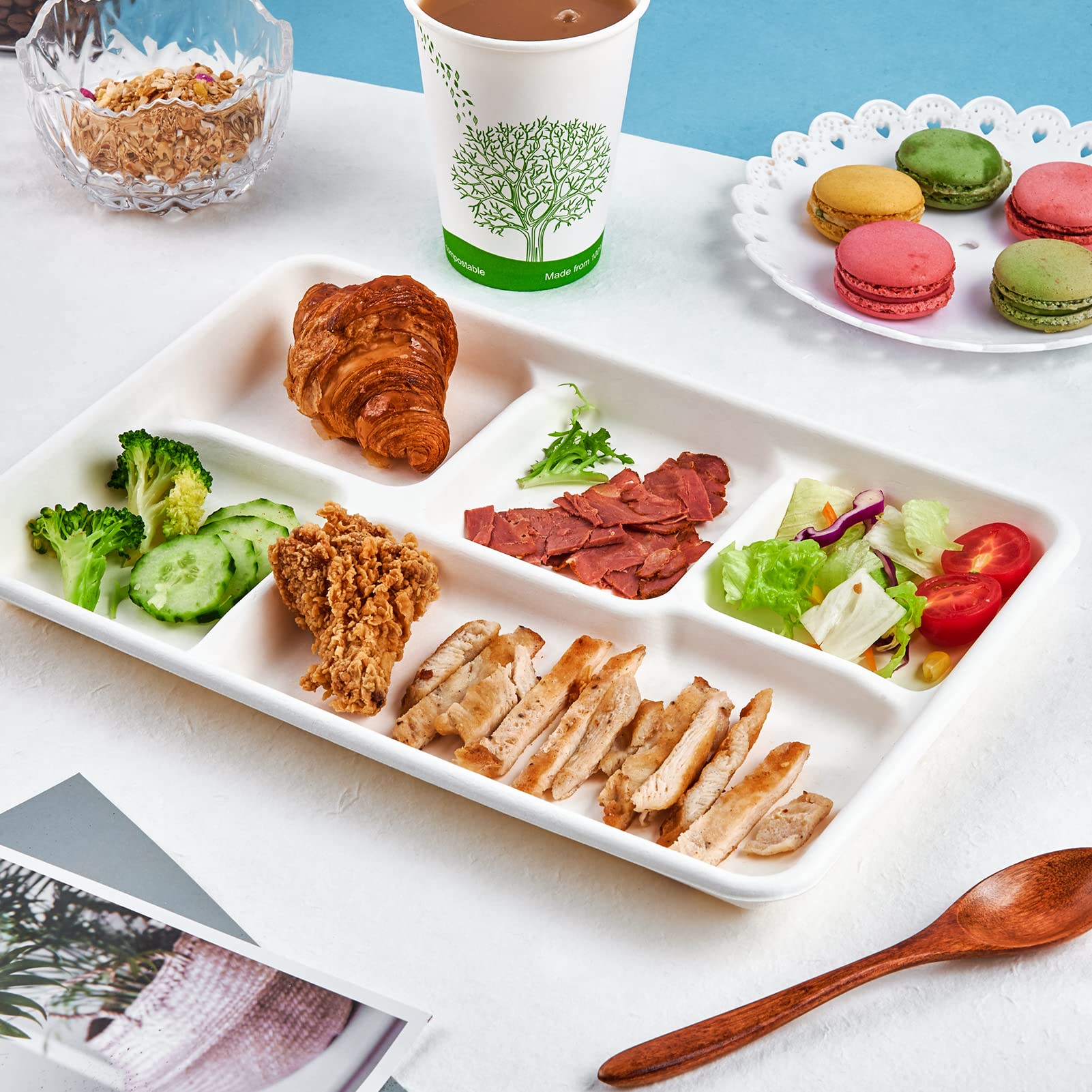 A white ECOlipak eco - friendly sugarcane pulp five - compartment meal tray holds a variety of foods, including croissant, broccoli, cucumber, fried chicken, sliced chicken, bacon, and salad. Alongside are a paper cup with a tree logo, macarons on a doily - edged plate, a bowl of granola, and a wooden spoon.