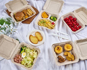 Assorted Ecolipak biodegradable meal containers with foods like burgers, pasta, salads, pastries and fruits, on a white cloth with cutlery and flowers.