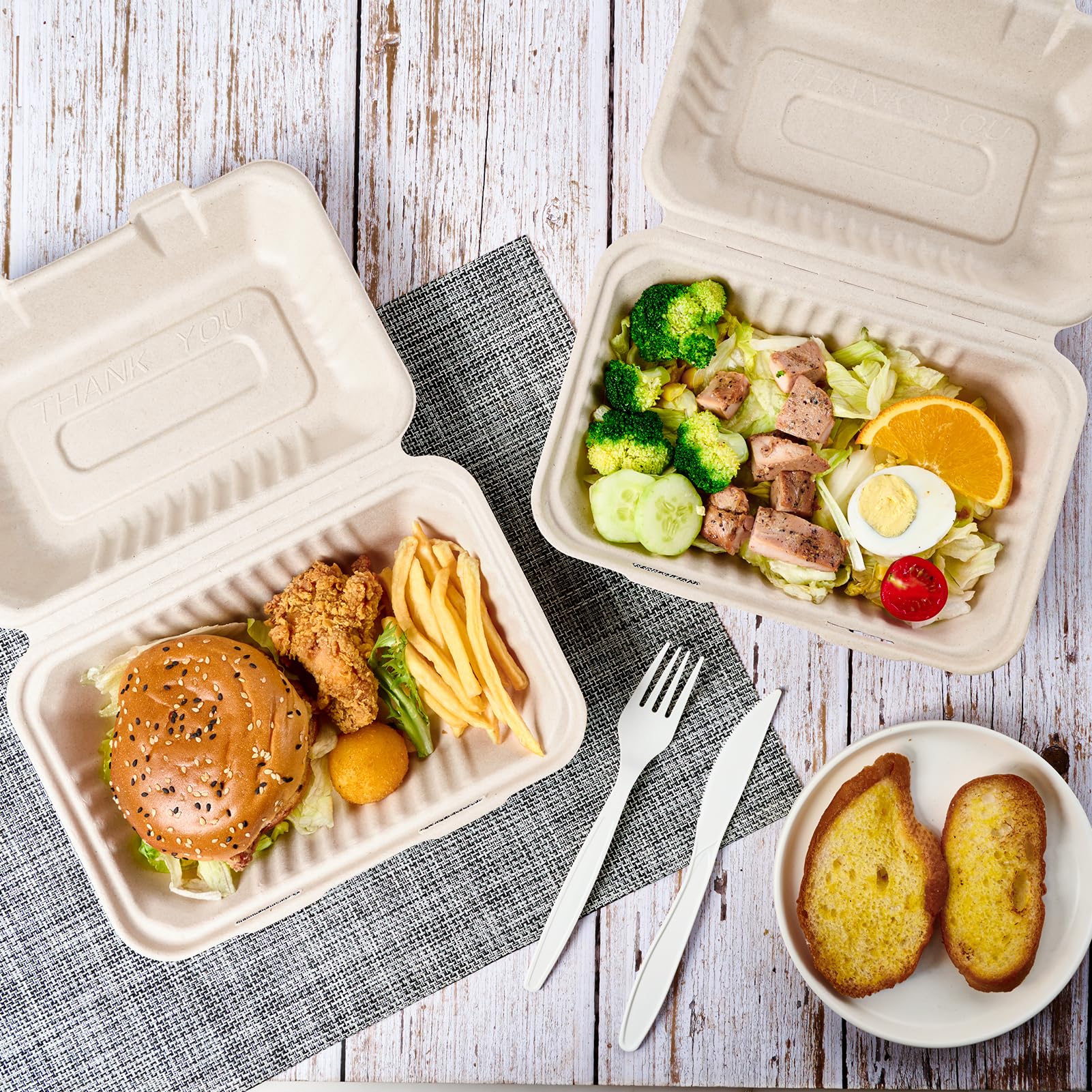 A to-go meal featuring a burger with fried chicken, fries, and a salad with broccoli and sliced meats, accompanied by garlic bread.
