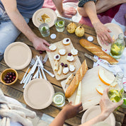 A picturesque picnic scene featuring plates of food, glasses of drinks, and hands reaching for snacks on a wooden table.