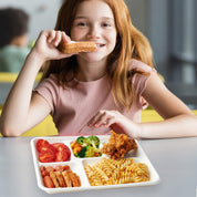 A child holds a snack while sitting at a table with a Compostable White Sugarcane 5 Compartment Plates tray featuring pasta, fried chicken, tomatoes, and mixed vegetables.