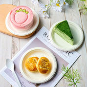 ECOlipak sugarcane pulp bowls hold a strawberry - shaped cake, matcha crepe, and sesame - topped pastries. A CPLA - material spoon, both made from eco - friendly materials, lies beside. The setting includes a magazine and flowers on a wooden surface.