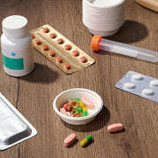 A variety of pills and capsules are spread on a wooden surface, including a bottle, blister packs, and a measuring syringe.