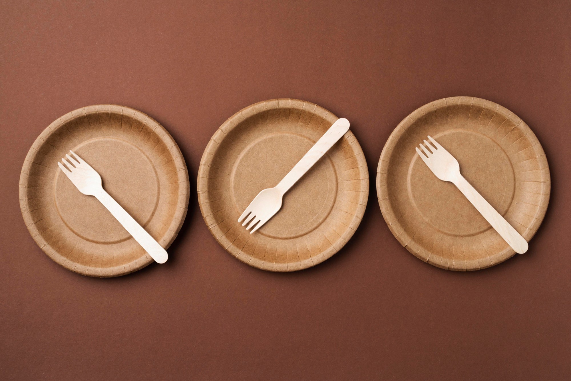 Three brown disposable plates arranged in a row, each with a  ECOLipak  wooden fork placed on top, set against a darker brown background.