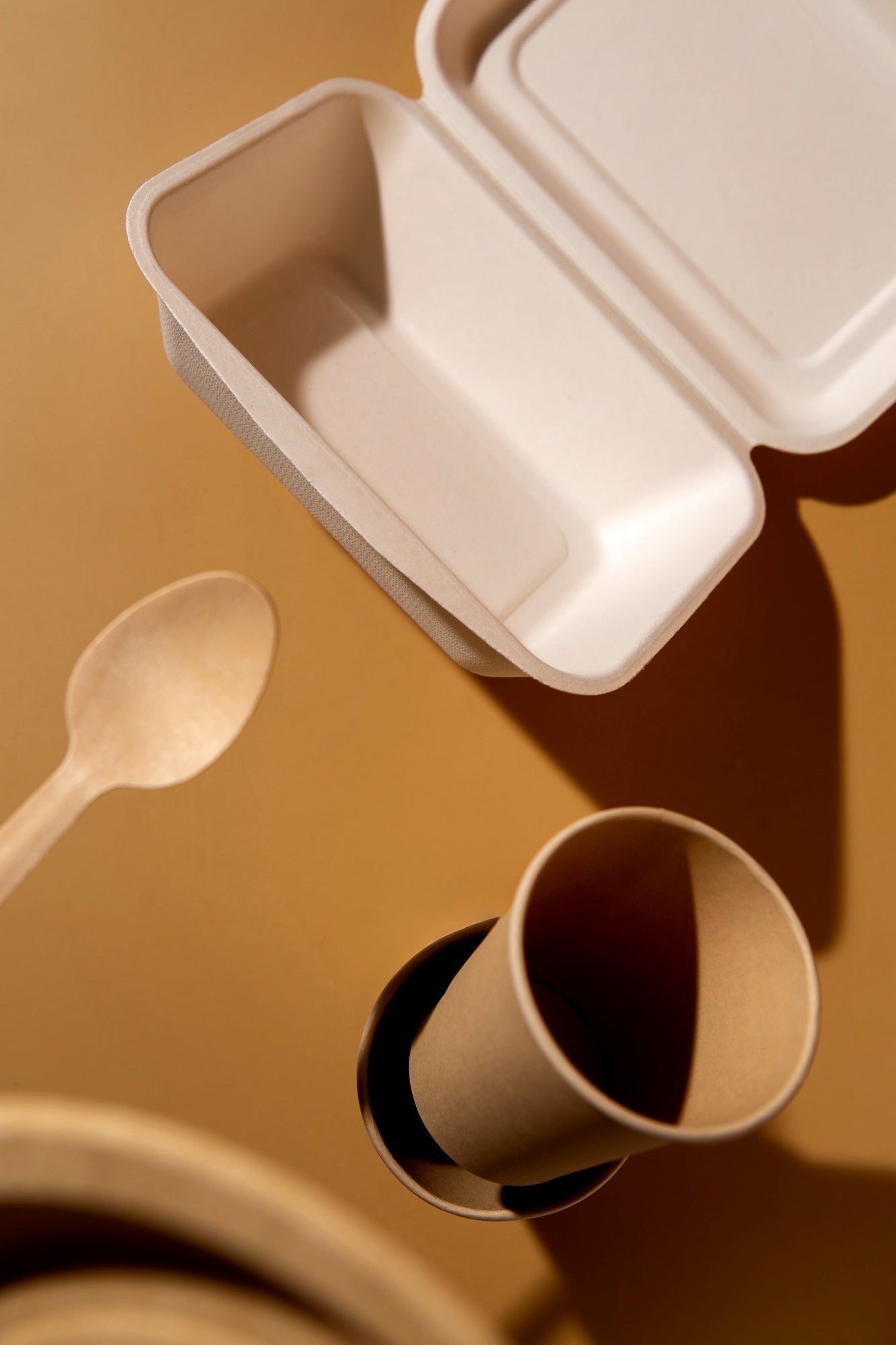 A ECOLipak white takeout box lies open on a gold surface, accompanied by a ECOLipak wooden spoon and a rolled paper cup.