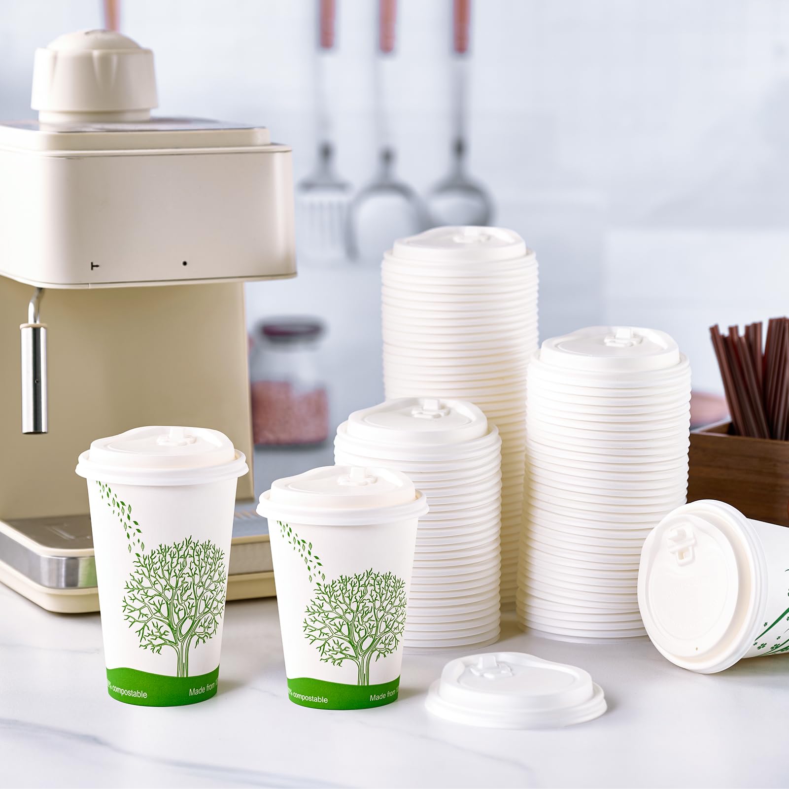 Two eco-friendly coffee cups with tree designs beside a stack of ECOLipak white lids, near a coffee machine and utensils in a bright kitchen.