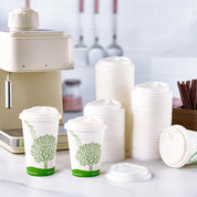Two eco-friendly coffee cups with tree designs beside a stack of ECOLipak white lids, near a coffee machine and utensils in a bright kitchen.