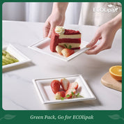 A hand lifts a slice of red velvet cake with strawberries, set on a square plate, near plates of kiwi and orange slices.